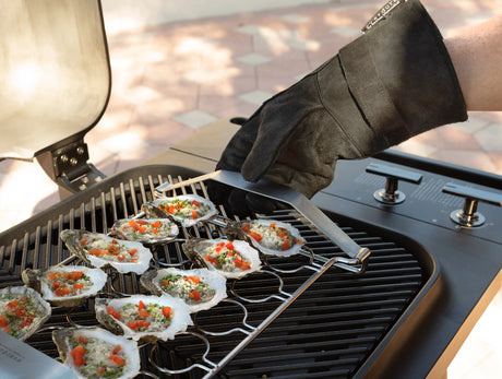 Person grilling oysters on a barbecue with a gloved hand using tongs.